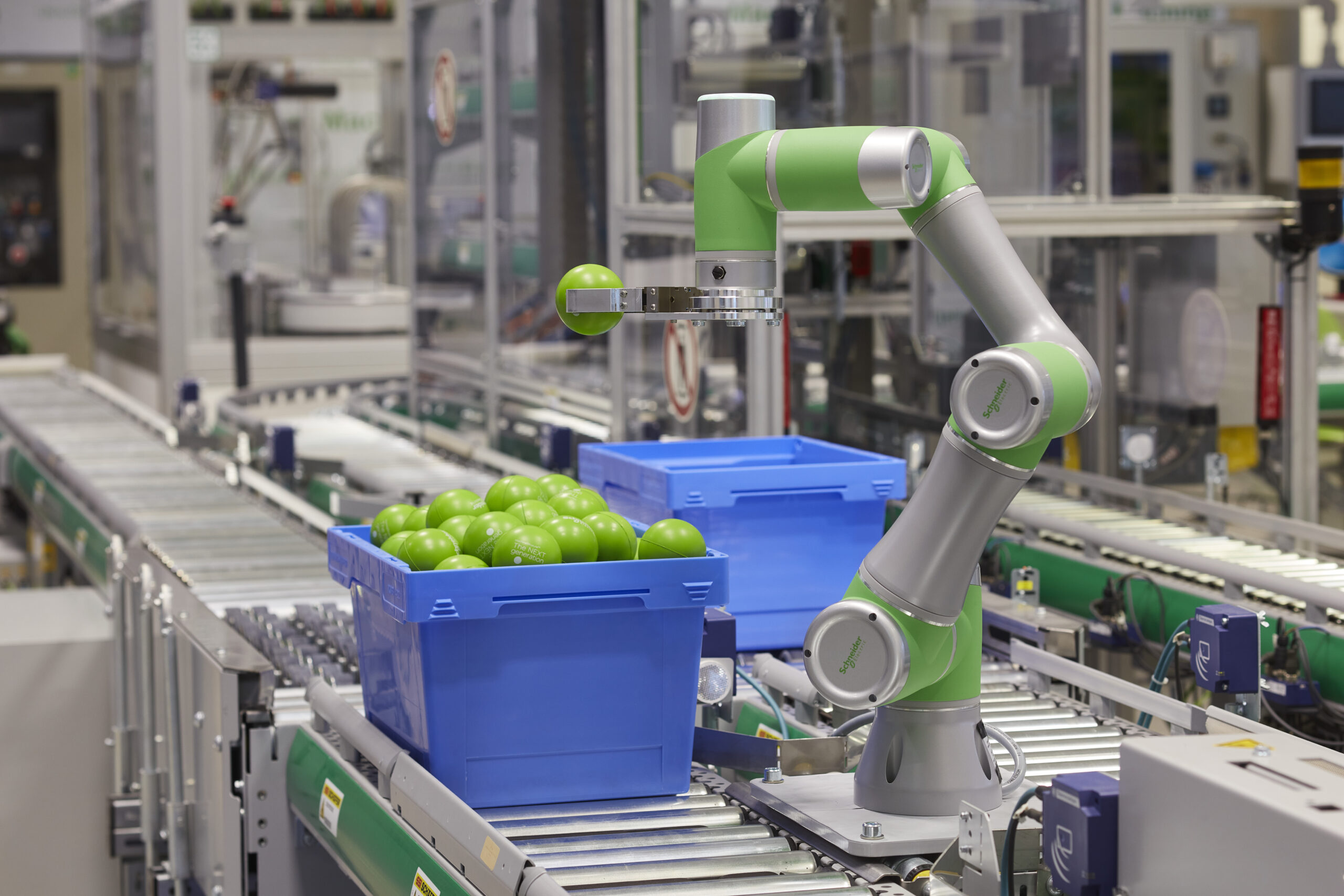 A robotic arm lifts one green plastic ball out of a bin full of many balls on a conveyor line in a factory.